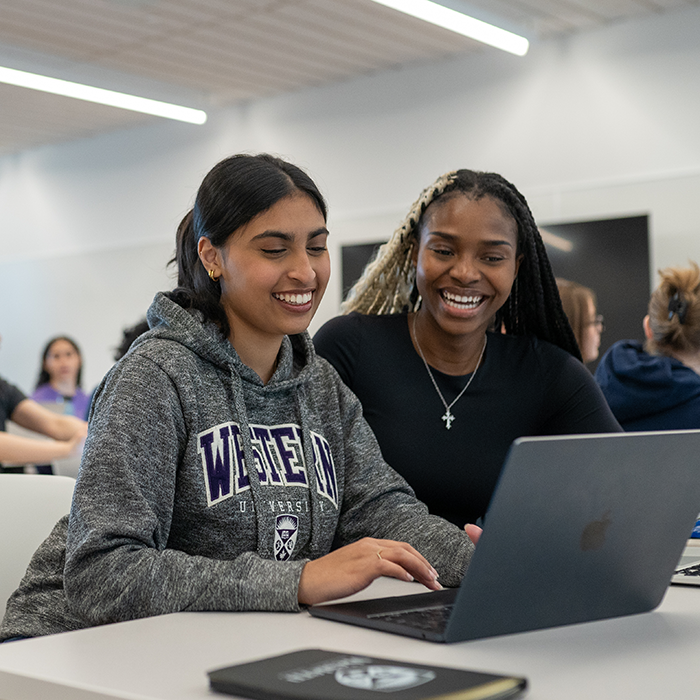 Two students laughing while working on a laptop in a classroom